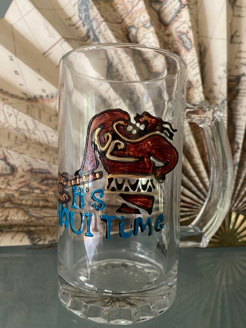 Puede incluir: Una jarra de cerveza de vidrio transparente con un dise&ntilde;o pintado a mano de Maui de la pel&iacute;cula Moana. El dise&ntilde;o est&aacute; en tonos de marr&oacute;n, dorado y azul. El texto "It's Maui Time" est&aacute; escrito en azul en la jarra.