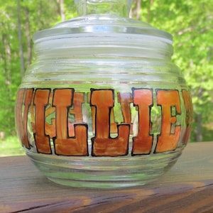 May include: A clear glass jar with a lid, decorated with the word "WILLIE" in orange and black paint.