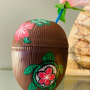 May include: A brown coconut-shaped cup with a green turtle and pink flowers painted on it. A pink flower with a blue straw is on top of the cup.