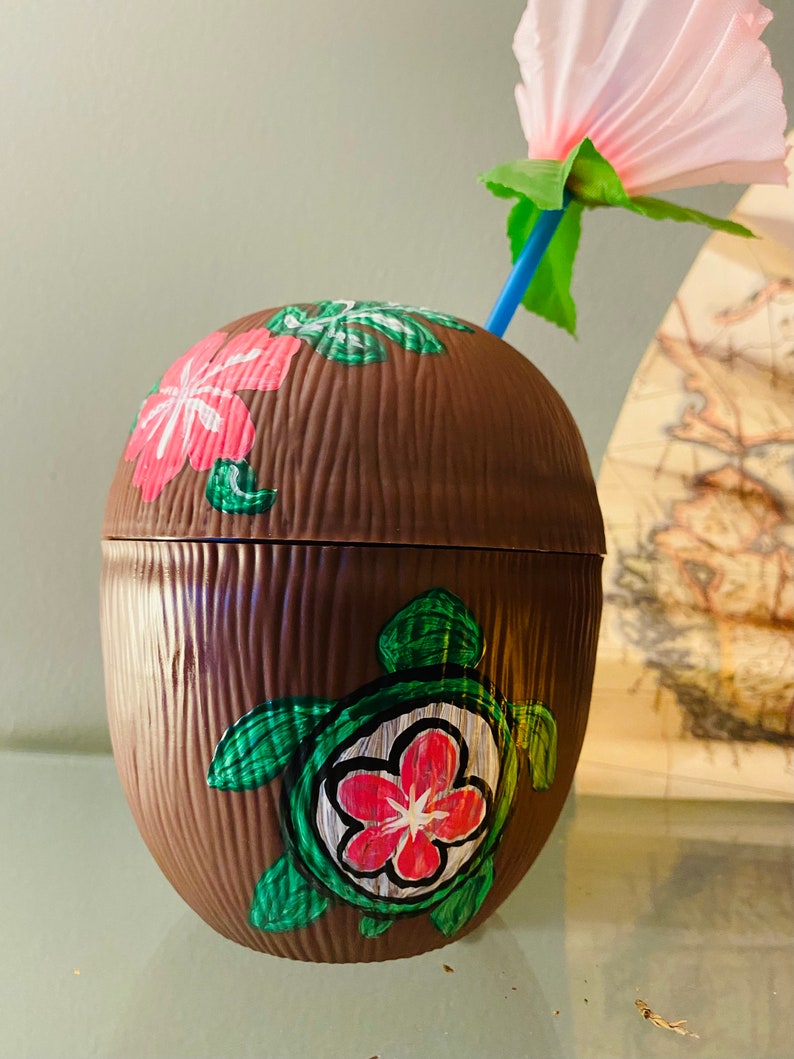 May include: A brown coconut-shaped cup with a green turtle and pink flowers painted on it. A pink flower with a blue straw is on top of the cup.