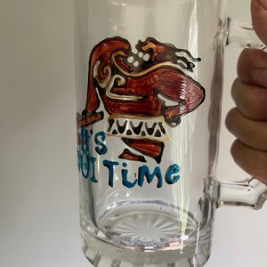 Puede incluir: Una taza de vidrio transparente con un dise&ntilde;o marr&oacute;n y dorado de una persona con una falda de estampado azul y blanco. El texto "It's Oui Time" est&aacute; escrito en azul en la taza.