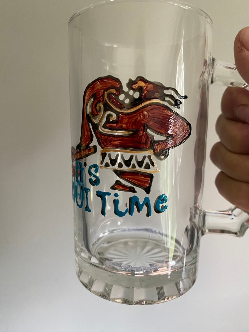 Puede incluir: Una taza de vidrio transparente con un dise&ntilde;o marr&oacute;n y dorado de una persona con una falda de estampado azul y blanco. El texto "It's Oui Time" est&aacute; escrito en azul en la taza.