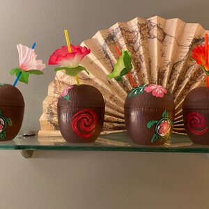 May include: Four brown coconut-shaped cups with colorful floral designs and straws. The cups are on a glass shelf in front of a decorative fan.