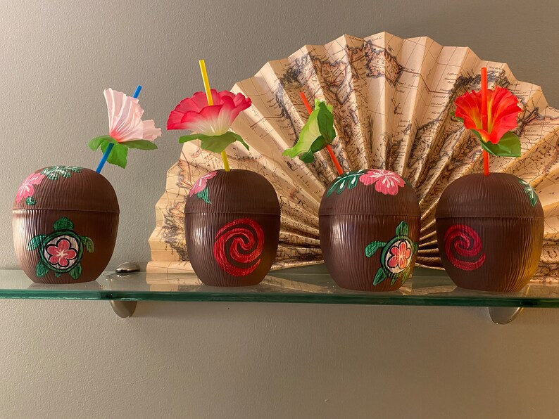 May include: Four brown coconut-shaped cups with colorful floral designs and straws. The cups are on a glass shelf in front of a decorative fan.