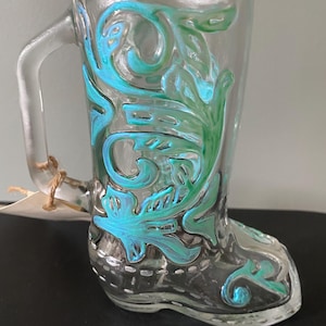 May include: A clear glass boot-shaped mug with a handle, decorated with teal and green floral patterns. The mug has a vintage appearance, with a small tag attached to the handle. The design is painted on the outside of the glass.