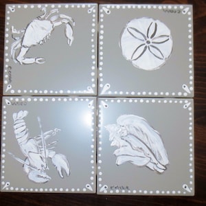Op de afbeelding: Vier vierkante keramische tegels met een grijze achtergrond, elk met een wit zeedierontwerp. De ontwerpen omvatten een krab, zanddollar, kreeft en zeeschelp. Elke tegel heeft een witte gestippelde rand en de handtekening van de kunstenaar.