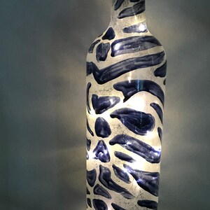 May include: A white glass bottle with a blue and white zebra stripe pattern. The bottle has a string of white lights inside.
