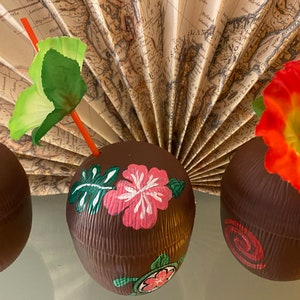 May include: Three brown coconut-shaped cups with painted tropical designs. One cup has a pink hibiscus flower, green leaves, and a turtle. Another cup has a pink swirl design. The third cup has a red hibiscus flower.