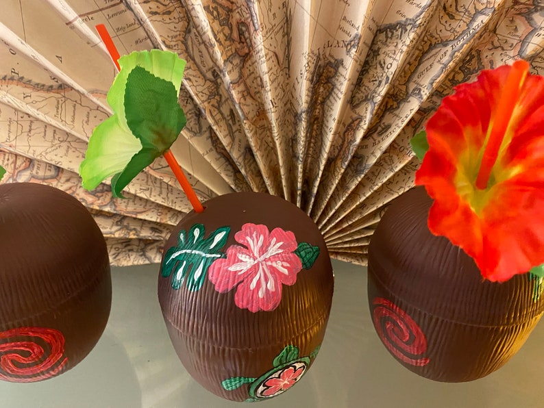 May include: Three brown coconut-shaped cups with painted tropical designs. One cup has a pink hibiscus flower, green leaves, and a turtle. Another cup has a pink swirl design. The third cup has a red hibiscus flower.