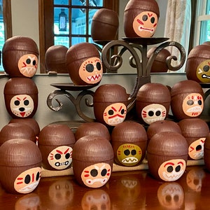 May include: A collection of brown coconut shell cups with painted faces. Each cup has a different facial expression and design. The cups are arranged in rows on a wooden surface.