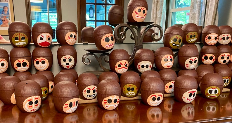 May include: A collection of brown coconut shell cups with painted faces. Each cup has a different facial expression and design. The cups are arranged in rows on a wooden surface.