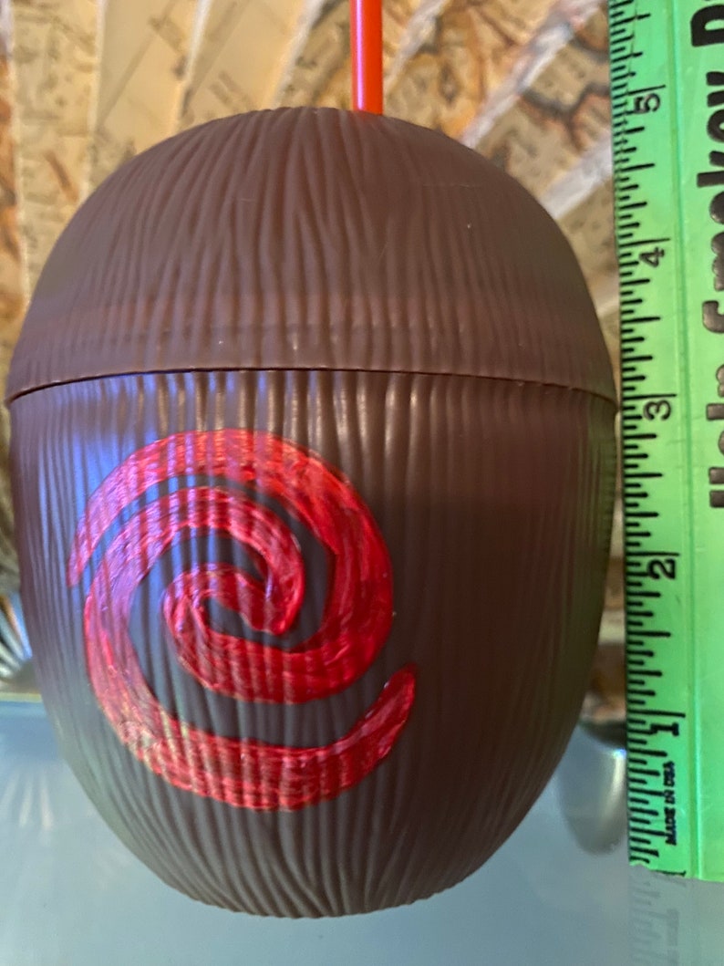 May include: Brown coconut-shaped cup with a red swirl design. The cup is about 6 inches tall and has a straw.