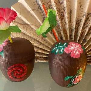 May include: Three brown coconut-shaped cups with painted tropical designs. The cups have a textured surface and are decorated with pink and white flowers, green leaves, and a turtle. One cup has a red swirl design.