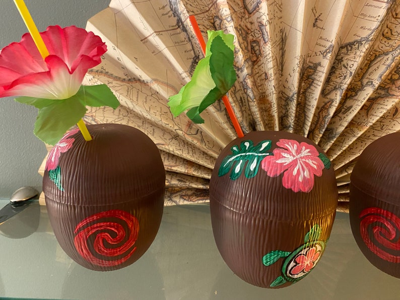 May include: Three brown coconut-shaped cups with painted tropical designs. The cups have a textured surface and are decorated with pink and white flowers, green leaves, and a turtle. One cup has a red swirl design.