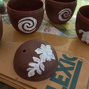 May include: Five brown coconut shell cups with white painted designs. The designs include a turtle, a spiral, and a leaf.