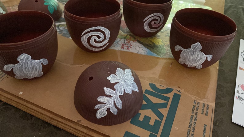 May include: Five brown coconut shell cups with white painted designs. The designs include a turtle, a spiral, and a leaf.