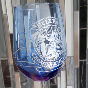 May include: A blue glass wine glass with a white design of a mermaid with the text "Shellback Crossing The 7 Seas".