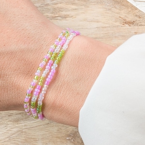 May include: A stack of three beaded bracelets in shades of pink, green, and clear. The bracelets are made of small, round beads strung together. The bracelets are worn on a wrist.