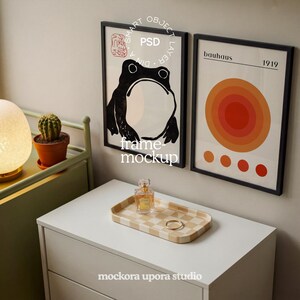 Könnte beinhalten: Zwei gerahmte Kunstdrucke mit schwarzen Rahmen hängen an einer Wand. Einer zeigt eine schwarze Froschillustration, der andere ein Bauhaus-Design mit orangefarbenen und roten Kreisen. Ein kariertes Tablett mit Parfüm und einem Ring steht auf einer weißen Kommode.