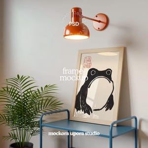 Puede incluir: Una lámpara de pared naranja con una pantalla redonda está encima de una impresión enmarcada de una rana en blanco y negro. La impresión está en un estante azul, con una planta en maceta a la izquierda. El texto "PSD" es visible en la lámpara.