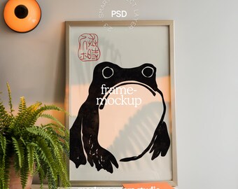 May include: A framed art print featuring a black and white frog illustration. The artwork has a minimalist style with the text "frame-mockup". A fern in a terracotta pot and a small orange book are in the foreground.