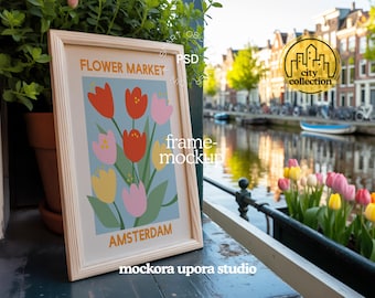 Op de afbeelding: Een ingelijste kunstdruk met een bloemenontwerp van rode, roze en gele tulpen op een lichtblauwe achtergrond. De woorden "FLOWER MARKET AMSTERDAM" staan op het kunstwerk. Het frame is van lichtgekleurd hout.