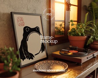 May include: A framed art print featuring a black frog illustration with Japanese text. A record player with a vinyl record sits on a wooden surface, alongside a decorative plate. Plants and a window are in the background.