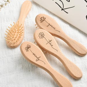 May include: A collection of wooden hairbrushes. Three brushes are engraved with names and floral designs. One brush has bristles. The brushes are light brown and sit on a white surface.