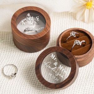Könnte beinhalten: Holzringboxen mit transparenten Deckeln, die Verlobungsringe zeigen. Eine Box hat die Initialen "A" und "F" mit einem floralen Design und den Worten "Apollo & Felicity". Eine andere Box hat den Text "Mr. & Mrs. Anderson 06.12.25".