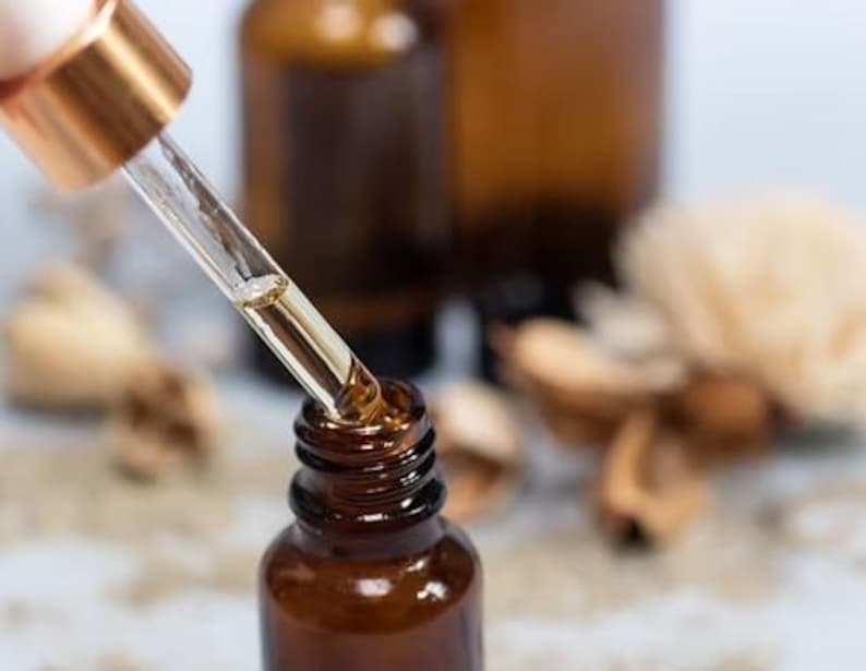 Op de afbeelding: Close-up van een bruine glazen fles met een druppelaar die een goudkleurige vloeistof afgeeft. De druppelaar heeft een goudkleurige bovenkant. Andere bruine flessen staan op de achtergrond, samen met wat lichtgekleurd, gedroogd plantmateriaal.