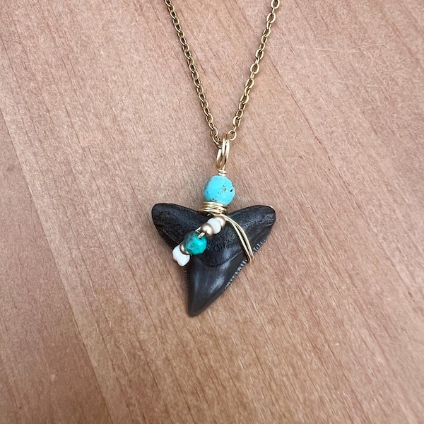 May include: A necklace featuring a dark gray fossilized shark tooth pendant wrapped in gold-toned wire. The pendant is accented with a single bright turquoise bead at the top and a small cluster of white and turquoise beads along the side wire wrap.