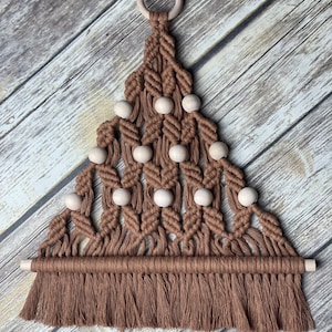 May include: A brown macrame wall hanging in the shape of a triangle with wooden beads and a wooden dowel at the bottom. The hanging has a fringe at the bottom.