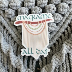 May include: A sticker with the words "macrame all day" in a decorative font. The sticker features a macrame design in shades of brown, green, and beige, set against a gray macrame background. The sticker is white with a decorative shape.