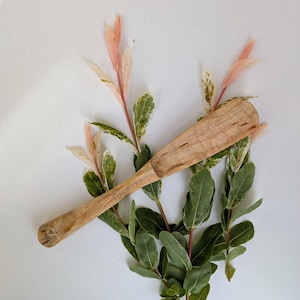 May include: A light brown wooden shoehorn with a carved handle and etched design. The shoehorn is displayed on a bed of green and pink-tipped leaves against a white background.