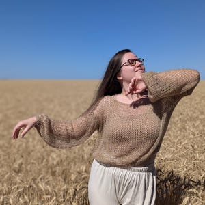 May include: A long-sleeved, loose-knit sweater in a metallic gold hue with a wide, rounded neckline and slightly puffed sleeves, paired with plain, light-colored trousers. The garment features a semi-sheer, open-weave texture throughout.