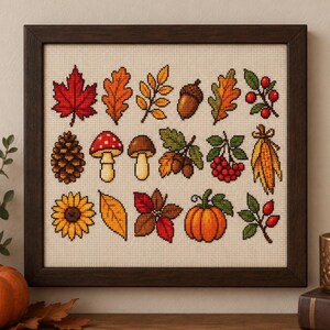 May include: A framed cross-stitch sampler on cream Aida cloth featuring a grid of autumn-themed motifs including a red maple leaf, oak leaves, an acorn, a pinecone, mushrooms, a sunflower, a pumpkin, berries, and a bundle of dried corn husks.