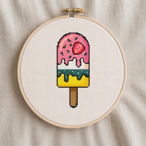 May include: A cross-stitch embroidery project in a round wooden hoop featuring a pixelated popsicle design. The popsicle has a pink top layer with a red strawberry slice, a white middle section, a teal layer, and a yellow base on a brown stick.