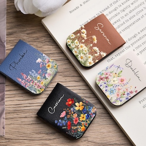 May include: Four square, leather-effect pocket mirrors featuring floral prints and personalised names. One blue mirror labelled Phoebe, one brown mirror labelled Serena, one cream mirror labelled Dahlia, and one black mirror labelled Cassian.