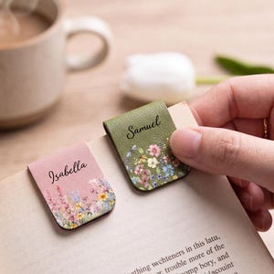 May include: Two magnetic bookmarks made of textured faux leather. One is pink with a floral print and the name Isabella in black script. The other is olive green with a floral print and the name Samuel in black script. Both are clipped to a book page.