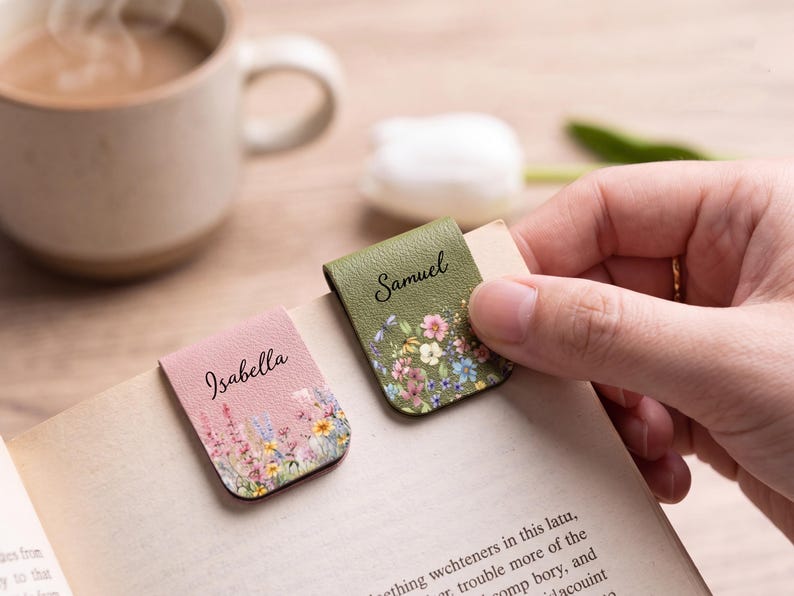 May include: Two magnetic bookmarks made of textured faux leather. One is pink with a floral print and the name Isabella in black script. The other is olive green with a floral print and the name Samuel in black script. Both are clipped to a book page.