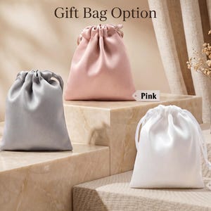 May include: Three satin drawstring gift bags in grey, pink, and white. Each bag features a smooth, lustrous finish and a matching ribbon closure at the top. Small rectangular tags labelled Grey, Pink, and White are placed next to each corresponding bag.