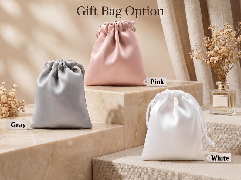 May include: Three satin drawstring gift bags in grey, pink, and white. Each bag features a smooth, lustrous finish and a matching ribbon closure at the top. Small rectangular tags labelled Grey, Pink, and White are placed next to each corresponding bag.