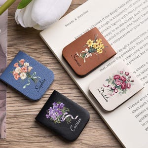 May include: Four leather bookmarks with floral designs and names. The bookmarks are in blue, brown, white, and black. Each features a different flower bouquet and a name: Sophia, Serena, Dahlia, and Cassian.