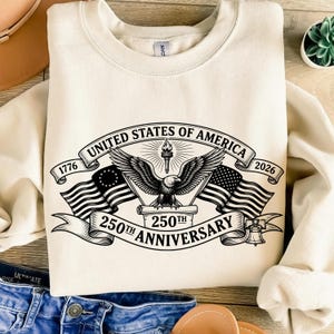 May include: Cream-colored sweatshirt featuring a black and white graphic commemorating the 250th anniversary of the United States of America. The design includes an eagle, flags, and the text "United States of America 250th Anniversary 1776-2026."