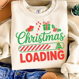 Könnte beinhalten: Cremefarbenes Sweatshirt mit den Worten "Christmas LOADING" in Grün und Rot. Das Design umfasst einen Weihnachtsstrumpf, eine Zuckerstange, ein Geschenk, Stechpalme und einen Weihnachtsbaum. Das Sweatshirt ist auf einer Holzoberfläche gefaltet.