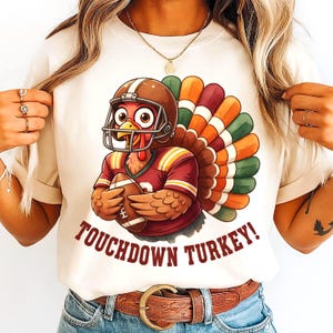 Pode incluir: Camiseta creme com um desenho animado de peru usando um capacete e uma camisa de futebol, segurando uma bola de futebol. O peru tem uma cauda colorida e o texto "TOUCHDOWN TURKEY!" em marrom. A camisa é usada por uma pessoa.