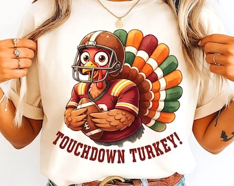 Könnte beinhalten: Cremefarbenes T-Shirt mit einer Cartoon-Truthahn, der einen Footballhelm und ein Trikot trägt und einen Football hält. Der Truthahn hat einen bunten Schwanz und den Text "TOUCHDOWN TURKEY!" in Braun. Das Shirt wird von einer Person getragen.