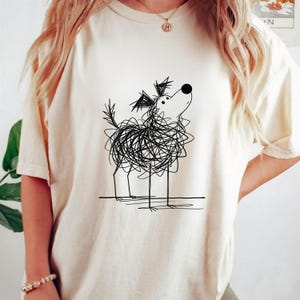 May include: Cream-colored t-shirt featuring a whimsical black line drawing of a dog. The dog's body is a scribble, with thin legs and a raised head. The design is centered on the shirt, offering a playful and artistic aesthetic.