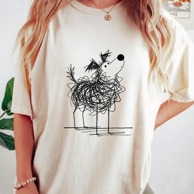 May include: Cream-colored t-shirt featuring a whimsical black line drawing of a dog. The dog's body is a scribble, with thin legs and a raised head. The design is centered on the shirt, offering a playful and artistic aesthetic.