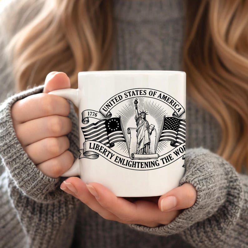 May include: White ceramic mug featuring a black and white graphic of the Statue of Liberty, American flags, and the text "United States of America" and "Liberty Enlightening the World". The mug has a curved handle.